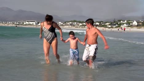 Madre-y-dos-niños-corriendo-a-través-del-agua-superficiales-en-la-playa,-Cape-Town