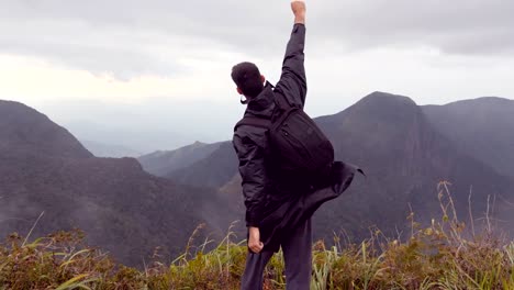 Young-male-hiker-in-raincoat-with-backpack-reaching-up-top-of-mountain-and-raised-hands.-Man-tourist-standing-on-the-edge-of-beautiful-canyon,-victoriously-outstretching-arms-up.-Rear-back-view