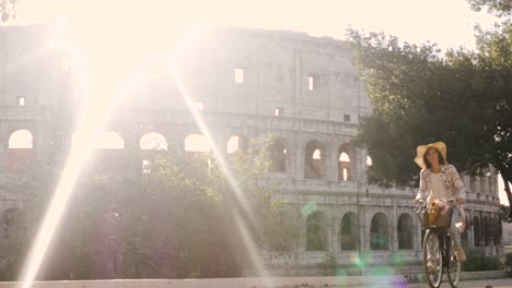 Schöne-junge-Frau-in-bunte-Mode-mit-Fahrrad-vor-Colosseum-in-Rom-bei-Sonnenuntergang-mit-Bäumen-glücklich-attraktives-Mädchen-mit-Strohhut-in-Colle-Oppio-Vorderansicht-Steadycam-dolly