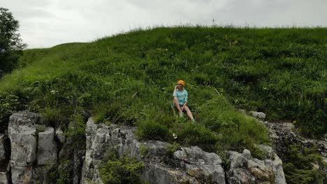 Young-hipster-woman-is-sitting-on-the-edge-on-the-grass-of-tall-wall-of-rock.-Aerial-view.-Drone-is-flying-slow-backward-from-model.-Establishing-revealing-shot