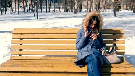 Woman-types-a-message-on-her-phone-sitting-on-the-bench-in-winter-city-park-in-sunny-day.