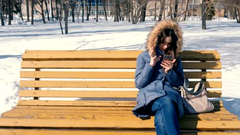 Frau-Typen-eine-Nachricht-auf-ihr-Handy-sitzen-auf-der-Bank-im-Stadtpark-Winter-im-sonnigen-Tag.