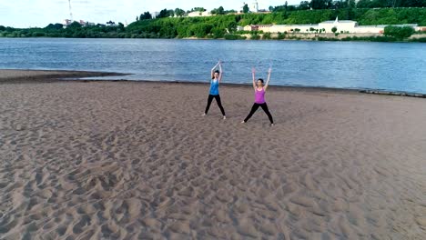 Zwei-Frau-Yoga-am-Sandstrand-am-Fluss-in-der-Stadt-zu-tun.-Blick-auf-die-schöne-Stadt-im-Sonnenaufgang.