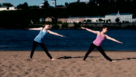 Zwei-Frau-Yoga-am-Sandstrand-am-Fluss-in-der-Stadt-zu-tun.-Blick-auf-die-schöne-Stadt-im-Sonnenaufgang.-Trikonasans-Utiha-darstellen.