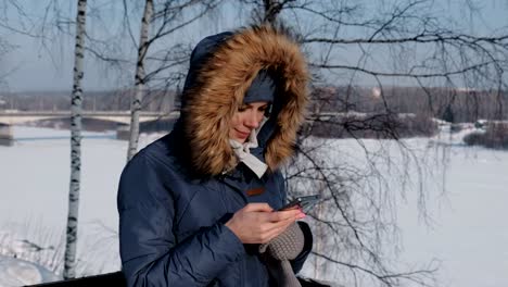 Woman-in-a-blue-down-jacket-with-a-fur-hood-writes-messaging-in-her-cellphone-in-a-winter-Park.