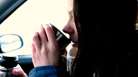 Junge-Brünette-Frau-trinkt-Tee-aus-einer-Thermosflasche-sitzen-in-einem-Auto-im-Winter.