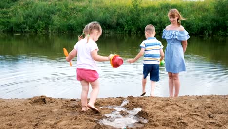 Los-niños-juegan-en-el-río-en-el-verano,-una-chica-y-un-chico-están-construyendo-una-corriente.-Mamá-cuida-a-los-bebés-durante-un-paseo