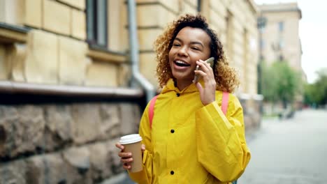 Ziemlich-Afroamerikanerin-ist-Kaffee-zum-Mitnehmen-und-im-Chat-auf-Handy-zu-Fuß-in-die-Stadt-im-Frühjahr-tragen-helle-Jacke.-Moderne-Technologie-und-Millennials-Konzept.