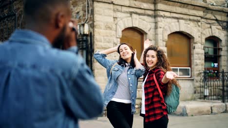 African-American-man-tourist-is-taking-pictures-of-his-female-friends-posing-for-camera-then-showing-them-photographs.-Tourism,-photography-and-youth-concept.