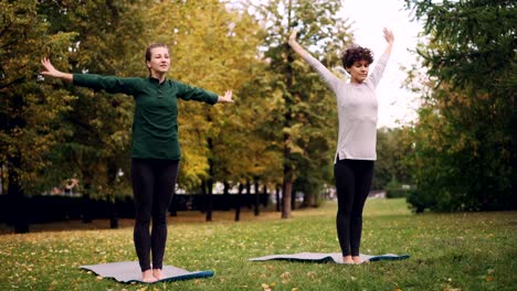 Two-good-looking-women-are-doing-yoga-in-park-on-mats-practising-asanas-and-breathing-fresh-air.-Individual-practice,-professional-teacher-and-nature-concept.