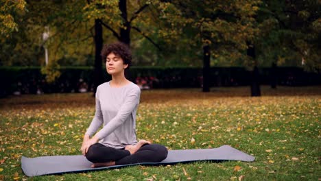 Chica-apuesta-en-deportes-ropa-es-torcer-su-cuerpo-sentado-en-posición-de-loto-y-luego-relajarse-con-ojos-cerrados-después-de-la-práctica-en-el-parque-en-día-de-otoño.