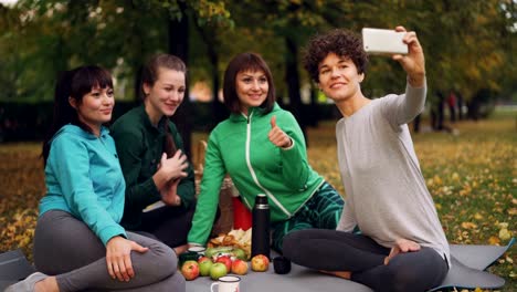 Attractive-young-ladies-yogini-are-taking-selfie-using-smartphone-during-picnic-in-park-in-autumn.-Girls-are-posing-and-smiling-having-fun-sitting-on-mats.