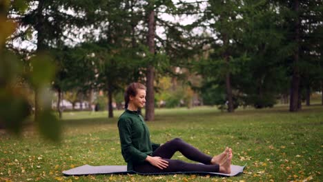 Sonriente-mujer-joven-está-ejerciendo-en-el-parque-haciendo-yoga-sentado-en-la-estera-de-hierba-verde-y-amarilla,-disfrutar-de-aire-fresco-y-saludable-actividad.-Concepto-de-juventud-y-recreación.