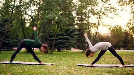 Estudiante-de-yoga-es-hacer-ejercicio-al-aire-libre-con-instructor-de-estiramientos-de-piernas-y-espalda-flexión-hacia-delante-de-pie-sobre-tapetes-en-el-prado-verde-y-amarillo-en-el-parque-de-la-ciudad.