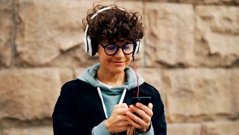 Pretty-girl-is-headphones-and-glasses-is-listening-to-music-and-touching-smartphone-screen-choosing-songs-standing-outdoors-in-city-with-stone-wall-in-background.