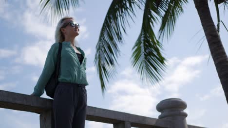 Young-happy-caucasian-woman-with-long-blonde-hair-in-sunglasses-and-green-shirt-standing-and-smiling-near-palm-tree-on-a-blue-sky-background.-Travel-concept