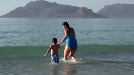 Mutter-und-junge-Jungen-spielen-im-seichten-Wasser-am-Strand,-Cape-Town