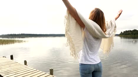 Young-woman-relaxes-on-lake-pier,-stands-arms-outstretched