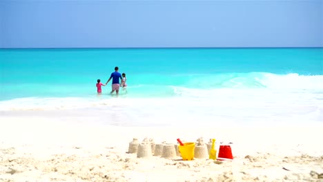 Sandburg-am-weißen-Strand-mit-Kunststoff-Kinderspielzeug-und-Familie-im-Meer-Hintergrund