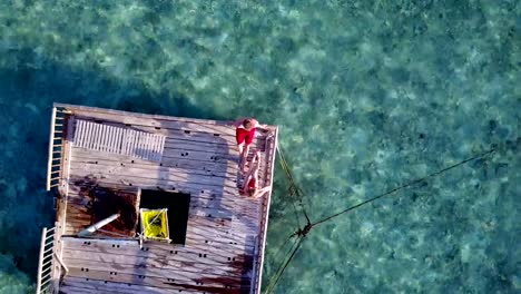 v03825-Aerial-flying-drone-view-of-Maldives-white-sandy-beach-2-people-young-couple-man-woman-relaxing-on-sunny-tropical-paradise-island-with-aqua-blue-sky-sea-water-ocean-4k-floating-pontoon-jetty-sunbathing-together