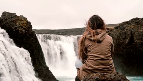 Rückansicht-der-junge-nachdenkliche-Frau-sitzt-auf-einem-Felsen-allein-und-auf-der-Suche-auf-mächtigen-Wasserfall-in-Island