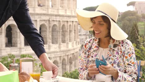 Turístico-de-la-hermosa-joven-con-smartphone-sentado-en-la-mesa-fuera-de-un-bar-restaurante-frente-al-Coliseo-de-Roma-proviene-de-café,-zumo-y-cornetto-el-camarero.-Elegante-vestido-con-sombrero-grande-y-bolsas-de-colores