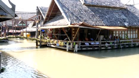 Mercado-flotante-de-Pattaya-y-casa-que-se-encuentra-en-el-mercado-flotante-de-Pattaya.-Personas-están-nadando-en-un-barco-en-un-río-marrón-a-Tailandia.-Poca-gente-a-lo-largo-de-la-orilla-y-montar-a-caballo-en-el-río-en-un-barco.