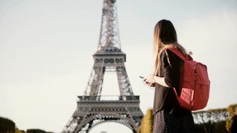 Young-beautiful-woman-with-backpack-standing-near-the-Eiffel-tower-and-using-the-smartphone,-browsing-the-Internet