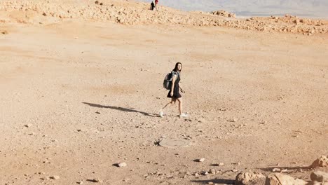 Woman-with-backpack-walks-to-desert-ruins.-Young-Caucasian-female-traveler-on-dry-sand,-rocks,-walls.-Masada-Israel-4K