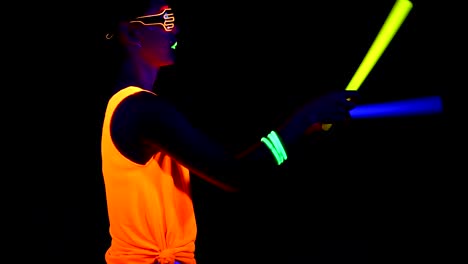 Woman-with-UV-face-paint,-glowing-clothing,-glowing-glasses,-bracelet-dancing-in-front-of-camera-holding-chemical-light,-half-body-side-shot.-Asian-woman.-.