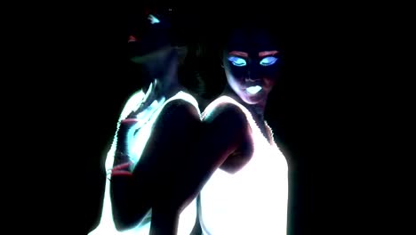 Women-with-UV-face-paint,-glowing-bracelets,-glowing-glasses,-clothing-dancing-back-against-each-other-in-front-of-camera,-Half-body-shot.-Caucasian-and-asian-woman.-Glitch-effect.-Women.