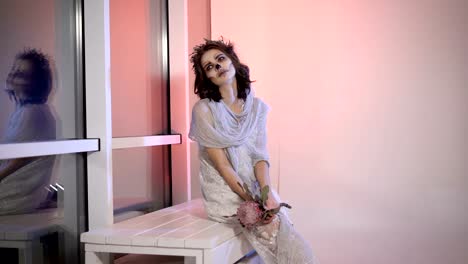 Close-up-of-a-young-girl-dressed-in-white-wedding-dress-with-creative-scary-and-creepy-halloween-make.-bizarre-appearance-dead-bride-is-sitting-on-the-bench-near-the-window