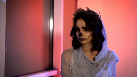 Close-up-of-face-a-young-girl-with-dead-mask-skull-art-on-her-.-bride-creative-scary-and-creepy-halloween-make-is-sitting-looking-at-the-window.-the-bench-the-pink-background