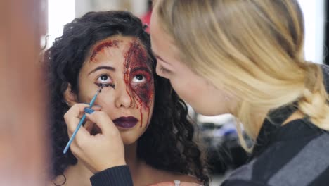 Make-up-artist-que-la-niña-halloween-hacer-upin-estudio.-Arte-de-cara-de-Halloween.-Mujer-se-aplica-a-maquillaje-profesional-en-la-cara-de-chica-española.-Pintura-de-guerra-de-sangre,-cicatrices-y-heridas.-4k