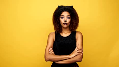 Joven-trastornado-a-mujer-africana-en-traje-de-Halloween-maquillaje-de-gato-y-orejas-buscando-cámara-aislada-sobre-amarillo-con