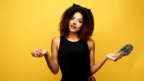 Carefree-african-lady-with-cat-make-up-holding-in-hands-candies-and-money,-trick-and-treat-isolated-over-yellow