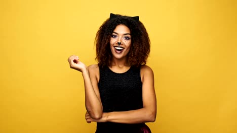 Señora-americana-afro-sorprendida-feliz-con-maquillaje-de-gato-y-orejas-sonriendo-a-cámara-aislada-sobre-pared-amarilla-con-murciélagos