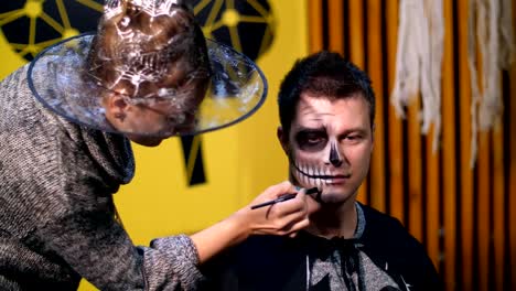 Halloween-party,-make-up-artist-draws-a-terrible-makeup-on-the-face-of-a-man-for-a-Halloween-party.-in-the-background,-the-scenery-in-the-style-of-Halloween-is-seen