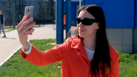 Portrait-of-happy-young-brunette-woman-in-sunglasses-makes-selfie-on-the-phone-the-beside-blue-building-on-the-street.