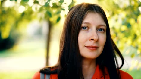 Close-up-portrait-of-charming-young-lady-smiling-and-looking-at-camera-standing-in-park-on-sunny-and-windy-day.-Healthy-lifestyle,-beautiful-nature-and-youth-concept.