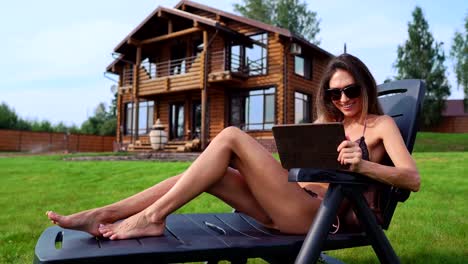 Eine-schöne-schlanke-Frau-liegt-auf-einer-Chaiselongue-in-einem-Badeanzug-mit-einem-Tablet-auf-dem-Hintergrund-eines-großen-Landhauses-in-Sonnenbrillen-und-sonnt-sich-in-der-Sonne,-die-Anzeige-von-Fotos-in-sozialen-Netzwerken