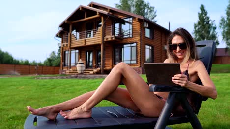 Una-hermosa-mujer-delgada-se-encuentra-en-una-hamaca-en-bañador-con-una-tableta-en-el-fondo-de-una-casa-rural-grande-en-gafas-de-sol-y-trabaja-remotamente-en-vacaciones