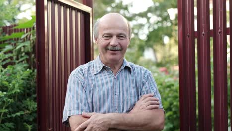 Friendly-caucasian-old-man-waving-hi-or-farewell,-isolated-outdoors-background-with-green-trees-and-fence