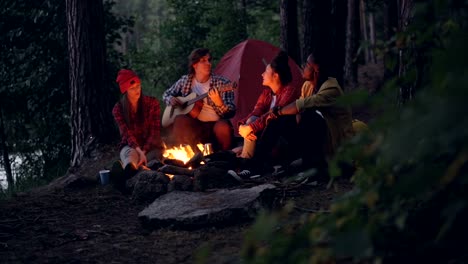 Girls-and-guys-friends-are-singing-songs-while-guitarist-is-playing-musical-instrument-sitting-near-fire-in-forest-and-enjoying-nature-and-music.-Tent-and-lake-are-visible.