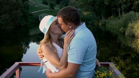 Ein-Liebespaar,-ein-Mann-und-eine-Frau-in-einem-weißen-Hut-auf-der-Brücke-im-Park-auf-dem-Hintergrund-von-einem-Teich-und-einer-wunderschönen-Landschaft,-umarmen-und-küssen.