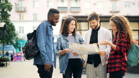 Grupo-multirracial-de-turistas-está-buscando-en-el-mapa-de-papel-de-pie-en-la-calle-en-ciudad-moderna-hablando-y-riendo.-Viaje,-navegación-y-concepto-de-juventud.