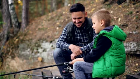 Bearded-young-man-is-fishing-with-cute-child-on-autumn-day,-boy-is-holding-rod-and-talking-to-father-learning-to-catch-fish.-People,-sharing-experience-and-family-concept.