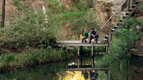 Loving-father-is-teaching-his-son-to-catch-fish-on-autumn-day,-people-are-sitting-on-wooden-pier-with-rods-and-talking-sharing-experience.-Recreation-and-family-concept.