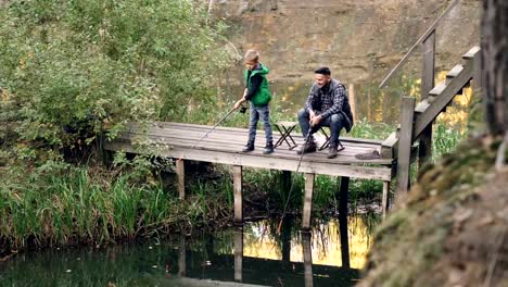 Fischers-Sohn-süße-Kind-ist-das-Fischen-mit-seinem-Vater-aus-hölzernen-Pier-holding-Stab-während-stolz-darauf,-dass-Eltern-sein-Kind-sucht-und-mit-ihm-zu-reden.-Elternschaft-und-Hobby-Konzept.