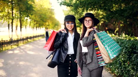 Retrato-de-adictos-a-las-compras-chicas-feliz-al-aire-libre-de-pie-con-los-bolsos-brillantes,-mirando-a-cámara-y-sonriendo-tocar-pelo.-Concepto-estilo-de-vida-comercial,-de-personas-y-de-la-juventud.